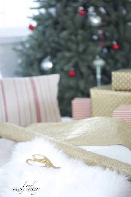 Gold scissors on top of a furry blanket with beige and red pillow and gifts wrapped in kraft paper in front of Christmas tree
