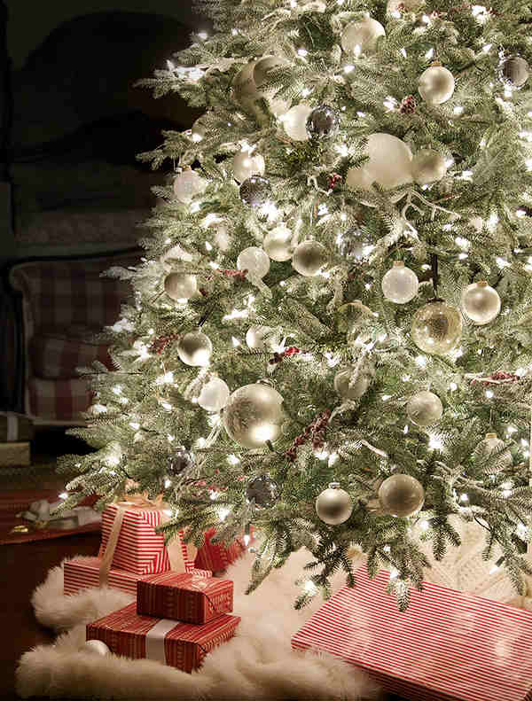 Closeup shot of a lit-up Christmas tree decorated with assorted white ornaments and a white fur tree skirt.