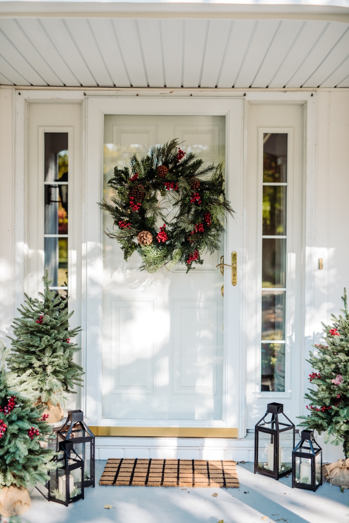 White front door decorated with Balsam Hill Outdoor Red Berry Pine Wreath, Balsam Fir Tabletop trees, and black lanterns