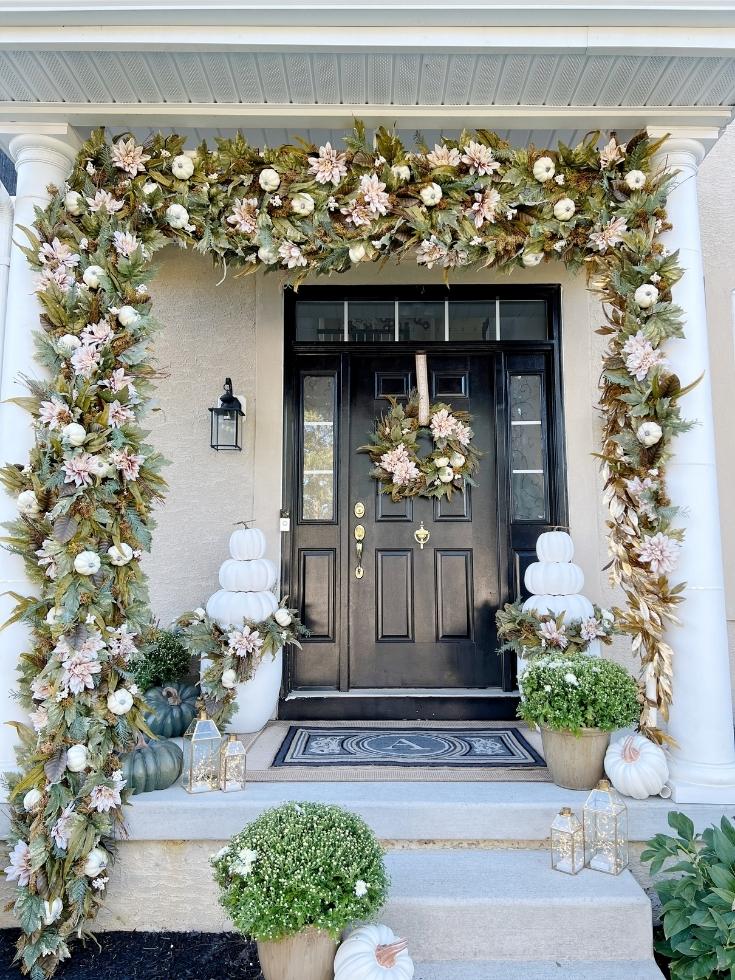A porch decorated with neutral colored fall decor such as dahlia wreath and garlands, white pumpkins, and brass fairy light lanterns
